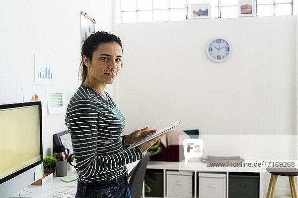 Lächelnde kreative Geschäftsfrau  die ein digitales Tablet hält  während sie im Büro steht