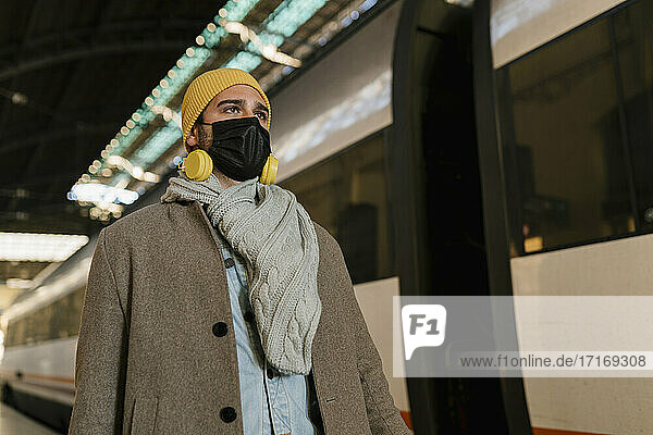 Mann mit Schutzmaske und Kopfhörern schaut weg  während er am Bahnhof am Zug steht