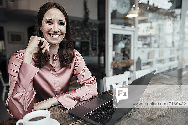 Lächelnde Unternehmerin mit Kaffeetasse und Laptop am Tisch in einem Cafe