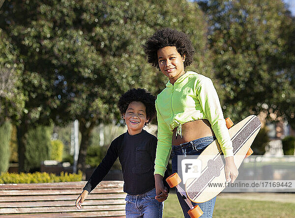 Mädchen hält Skateboard und die Hand ihres Bruders beim Spaziergang im Park