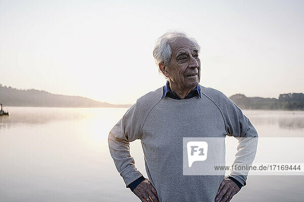 Älterer Mann mit Hand an der Hüfte  der wegschaut  während er am See steht