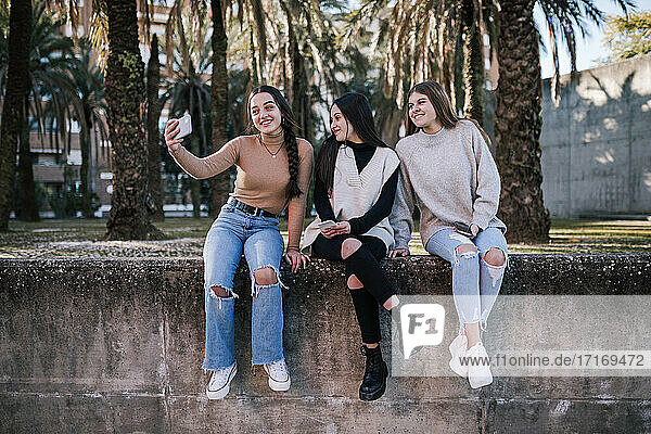 Smiling teenage girl taking selfie with friends while sitting on retaining wall in park