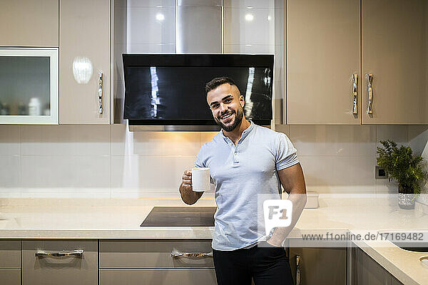 Handsome young man with hands in pockets holding coffee cup while standing in kitchen