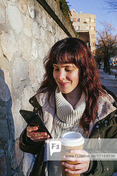 Lächelnde junge Frau  die ein Smartphone benutzt und einen wiederverwendbaren Becher an einer Steinmauer hält