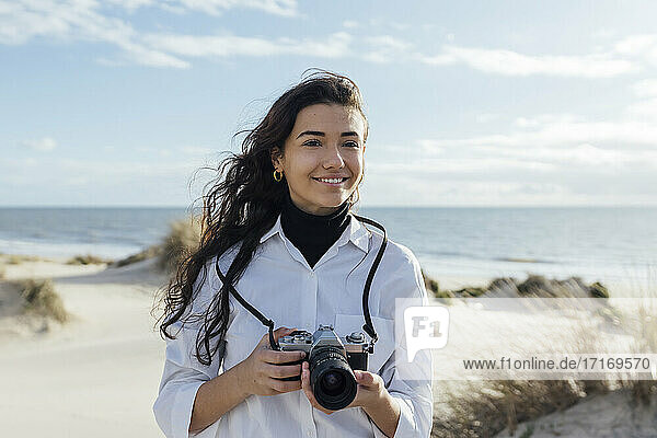 Beautiful smiling woman with camera at beach against cloudy sky