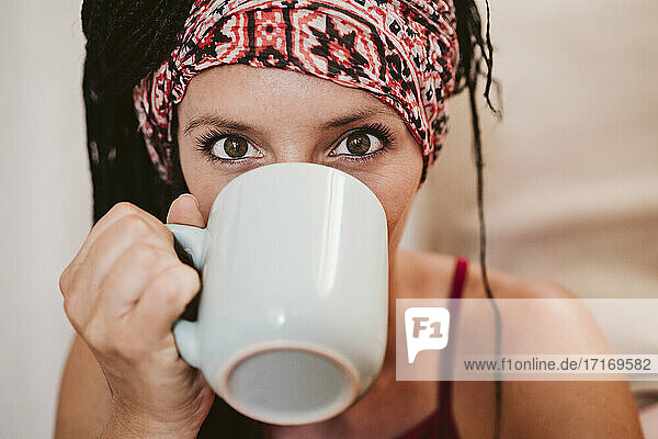 Mittlere erwachsene Frau mit Kopftuch trinkt Kaffee,  während sie zu Hause sitzt