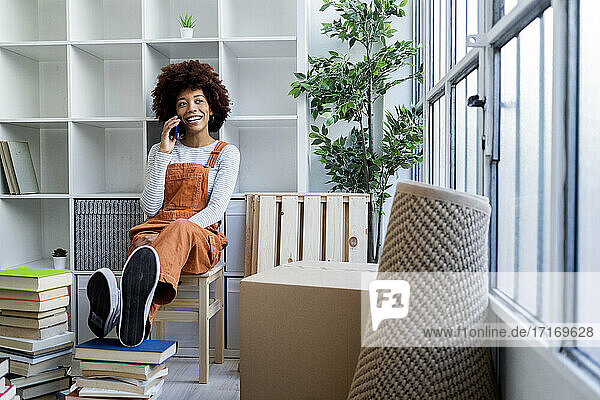 Afro-Frau  die mit ihrem Smartphone spricht  während sie an einem Bücherregal in ihrer neuen Wohnung sitzt