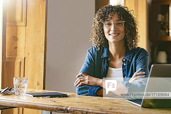 Happy young businesswoman with arms crossed by laptop at home office