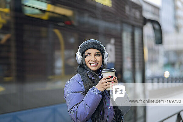 Smiling young woman with headphones holding coffee cup in city