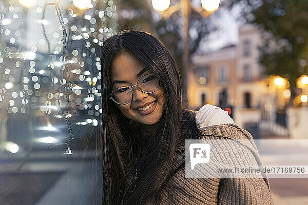 Happy young woman leaning on illuminated glass wall at dusk