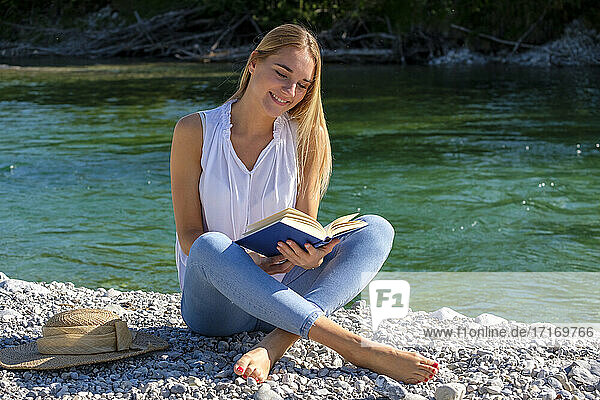 Lächelnde blonde Frau sitzt im Schneidersitz und liest ein Buch am Fluss