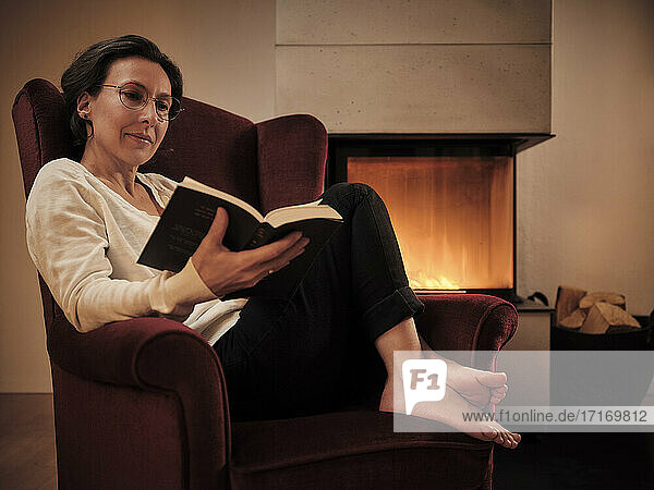 Reife Frau  die ein Buch liest  während sie zu Hause auf einem Sessel am Kamin sitzt