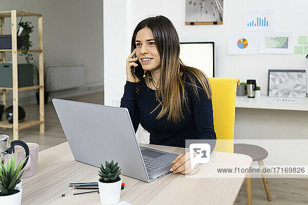 Smiling female freelancer on phone call while sitting by laptop at home