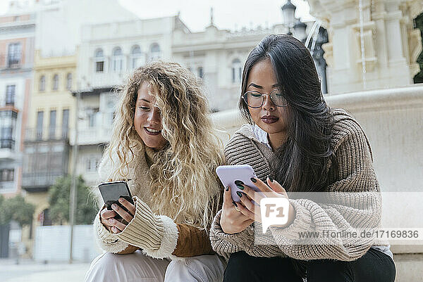 Multi-ethnic female friends using smart phone while sitting against fountain