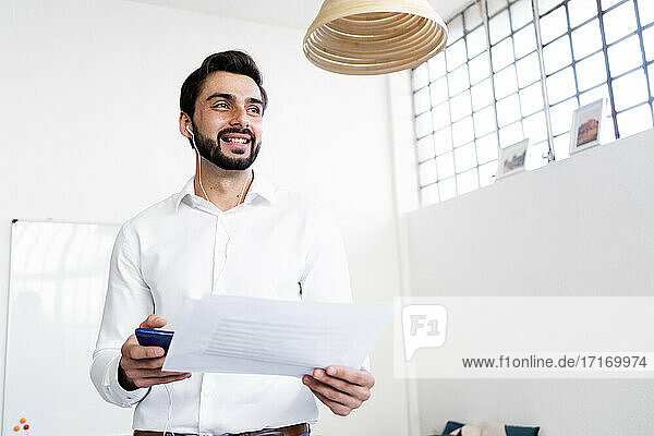Lächelnder Geschäftsmann mit Papierdokument bei einem Telefongespräch über In-Ear-Kopfhörer am Arbeitsplatz