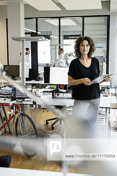 Fachkraft  die ein digitales Tablet benutzt  während sie mit einem Kollegen im Hintergrund in einem Großraumbüro steht