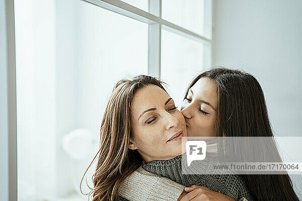 Tochter küsst Mutter auf die Wange am Fenster zu Hause