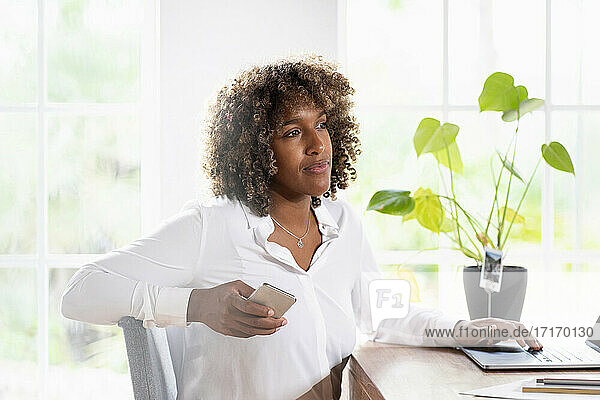 Thoughtful businesswoman with laptop and mobile phone looking away while sitting at home office