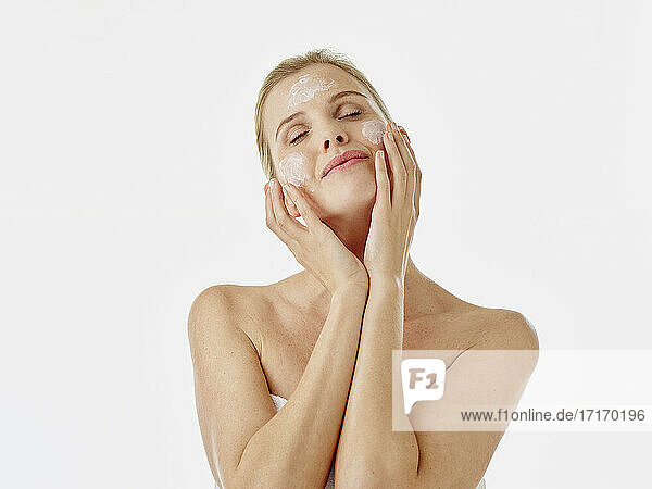 Woman wrapped in towel applying face cream while standing against white background