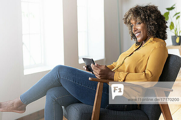 Lächelnde Frau mit digitalem Tablet  die wegschaut  während sie zu Hause auf einem Sessel sitzt