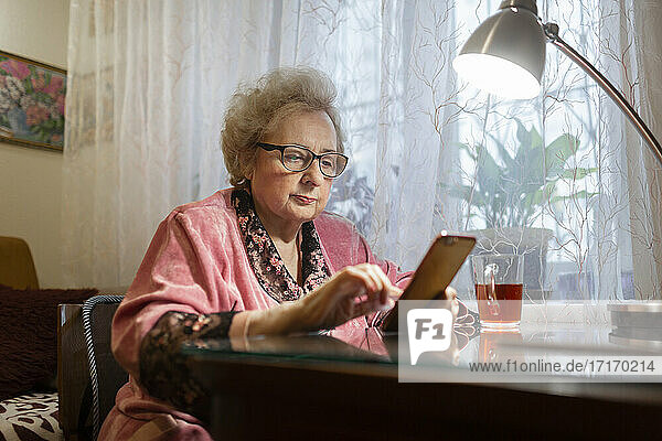Ältere Frau benutzt ihr Smartphone unter einer elektrischen Lampe auf dem Wohnzimmertisch