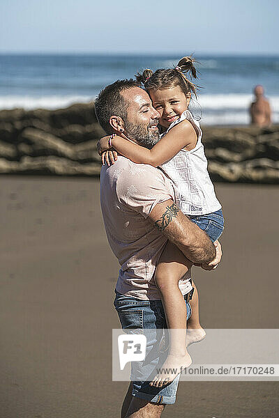 Smiling daughter embracing father at beach during sunny day