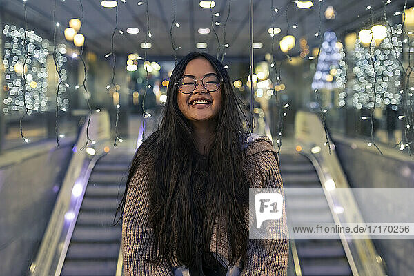 Cheerful young woman standing against illuminated glass wall in shopping mall