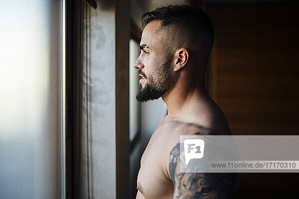 Shirtless man with tattoo looking through window at home