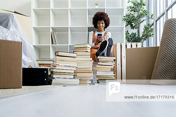 Afro-Frau  die ihr Smartphone benutzt  während sie in ihrem neuen Zuhause am Bücherregal sitzt