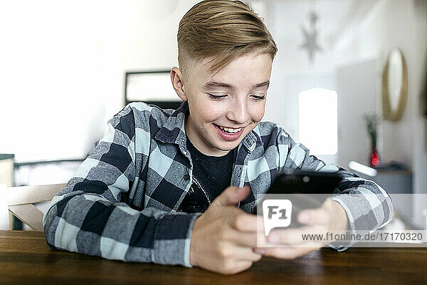 Lächelnder Junge  der ein Smartphone benutzt  während er am Tisch im Wohnzimmer sitzt