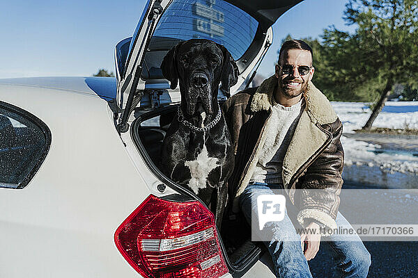 Junger Mann mit Hund sitzt im Kofferraum eines Autos an einem sonnigen Tag