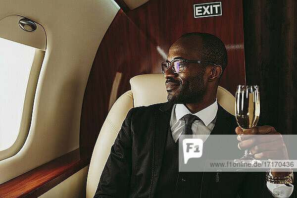 Lächelnder männlicher Unternehmer mit Champagner in der Hand  der durch das Fenster eines Privatjets schaut