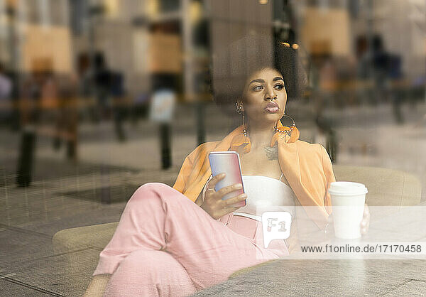 Afro woman with smart phone and coffee cup seen through glass at cafe