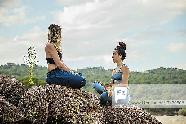 Flexible female instructor and young woman practicing yoga on rock against sky