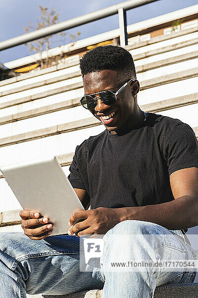 Lächelnder junger Mann  der ein digitales Tablet benutzt  während er auf den Stufen eines sonnigen Tages sitzt