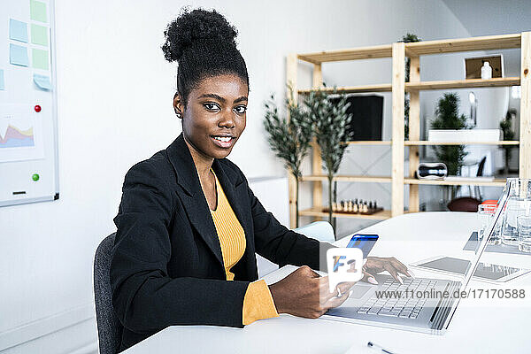Young female Afro entrepreneur sitting with laptop and smart phone at desk in office