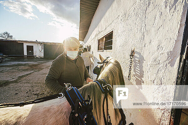Expertise wearing face mask standing by horse at stable