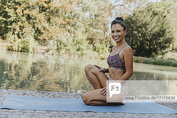 Female athlete smiling while sitting on exercise mat by lake at park