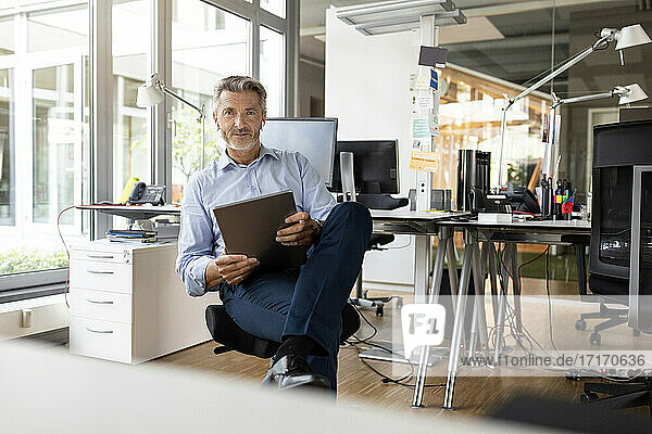 Confident entrepreneur with digital tablet sitting at open plan office
