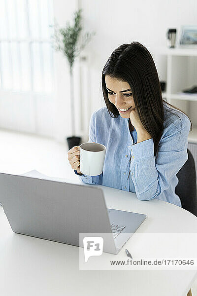 Berufstätige Frau trinkt Kaffee  während sie im Büro sitzt und einen Laptop benutzt