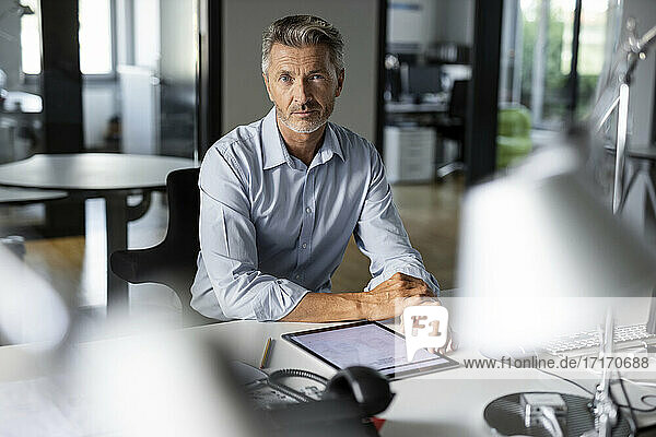 Mature confident businessman with digital tablet staring while sitting at office
