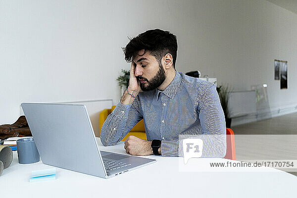 Müder Geschäftsmann mit Blick auf den Laptop im Kreativbüro