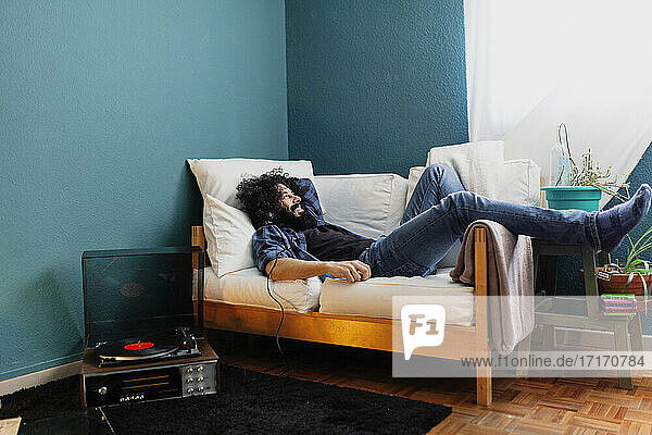 Bearded man listening music through turntable while lying on sofa at home