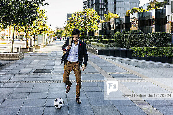 Businessman playing with soccer ball while running on footpath