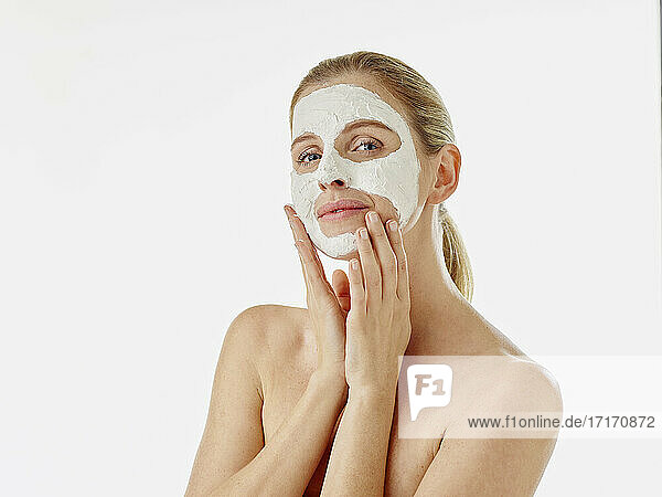 Young woman staring while applying face mask standing against white background
