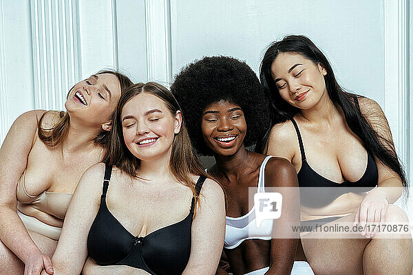 Glückliche multiethnische Gruppe von Modellen in Dessous mit geschlossenen Augen  die an einer weißen Wand sitzen