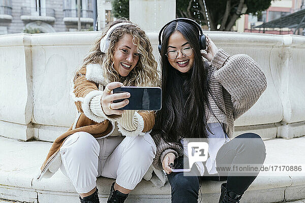 Smiling female friends taking selfie wearing headphones while sitting against fountain