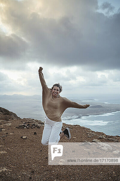 Carefree young woman jumping with arms outstretched on mountain against Famara Beach  Lanzarote  Spain