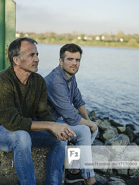 Father and son contemplating while sitting on rock against river
