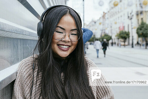 Smiling woman with headphones leaning on metal wall in city